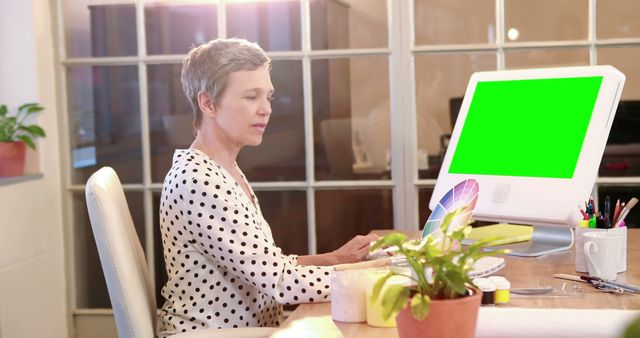 Senior woman uses computer with green screen in a modern office environment. This can be used for illustrating senior workers adapting to technology, office cleanliness, or promotional materials for office productivity.