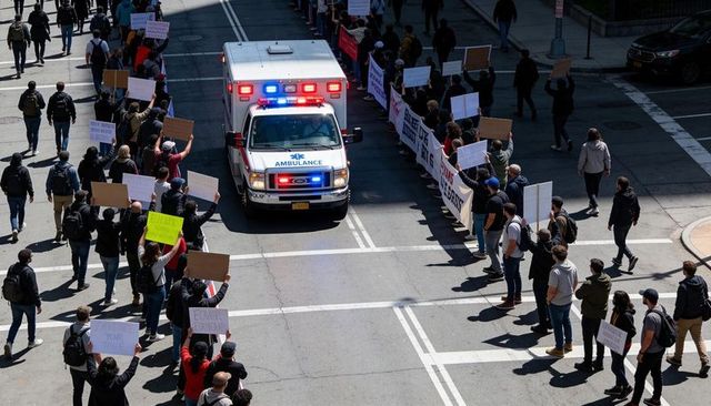 Ambulance driving through downtown protest with flashing lights, marching crowd, placards