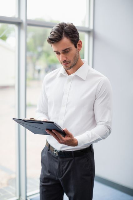 Businessman standing near large window, reviewing documents on clipboard. Ideal for use in business, corporate, and professional contexts, such as websites, presentations, and marketing materials.