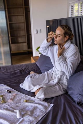 Woman in white bathrobe using facial rollers on bed morning skincare routine