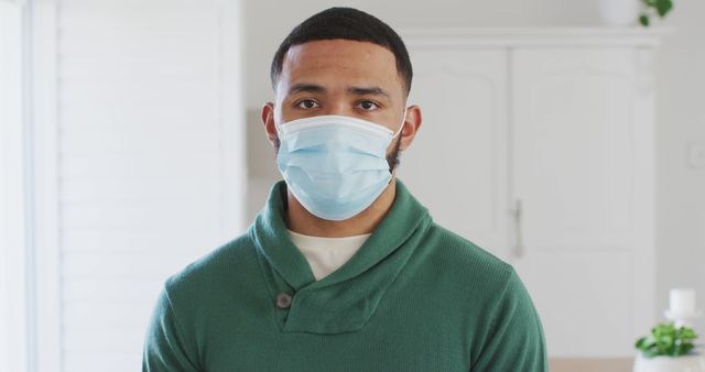 Young man indoors wearing a protective face mask looking at the camera. The image depicts a health-conscious young man in a casual green sweater taking precautions against COVID-19. Perfect for use in articles, health awareness campaigns, brochures, and social media posts emphasizing the importance of masking and safety during the pandemic.
