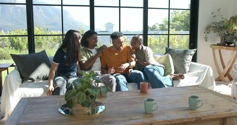African american friends laughing together on sunlit living room sofa with coffee mugs
