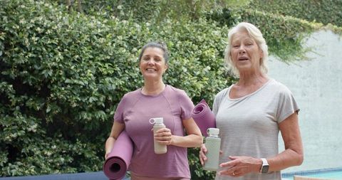 Mature women carrying yoga mats and water bottles, smiling poolside in lush backyard