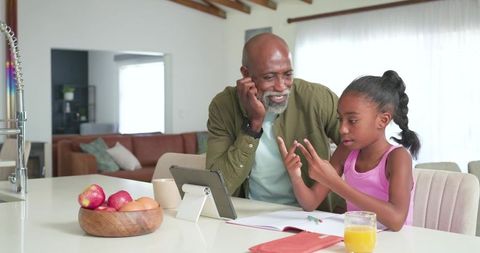 Grandfather tutoring granddaughter at kitchen island with tablet, notebook and orange juice