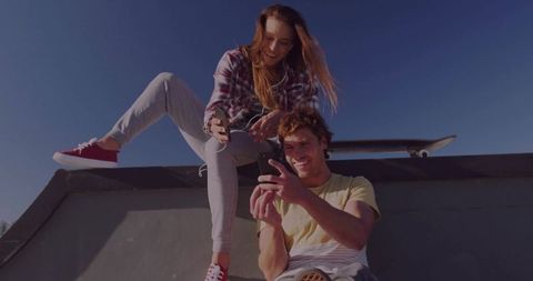 Young couple scrolling phones on skate ramp with skateboard and earbuds, casual lifestyle