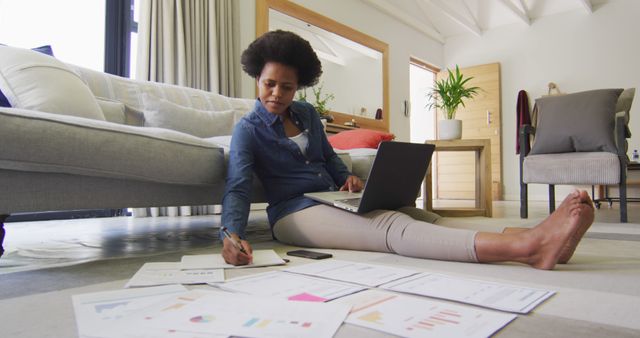 Young professional woman working remotely from home. She is sitting on the floor in a living room, analyzing documents spread around her while using a laptop. Ideal for business, remote work, freelance, home office, and productivity themes.