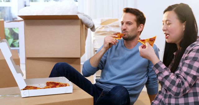 Man and woman taking a break from unpacking surrounded by moving boxes, sharing a slice of pizza. Perfect for depicting new beginnings, home relocation, or casual domestic life moments.