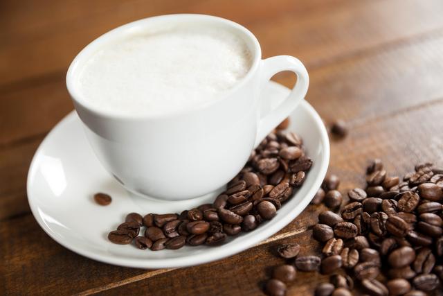 Cup of frothy coffee placed on a white saucer with coffee beans scattered on a wooden table. Ideal for use in breakfast advertisements, coffee shop promotions, or articles related to beverages and coffee culture.