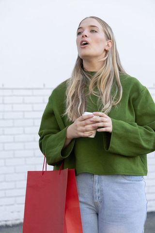 Young woman holding coffee and red shopping bag standing against white brick wall outdoors
