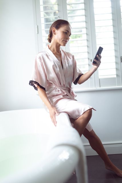 Woman using mobile phone while sitting on the edge of bathtub in bathroom at home