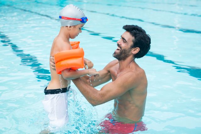 Father and son enjoying time together in a swimming pool at a leisure center. The father is holding his son, who is wearing floaties and a swim cap, ensuring his safety while they play. This image can be used for promoting family activities, swimming lessons, leisure centers, and summer fun.