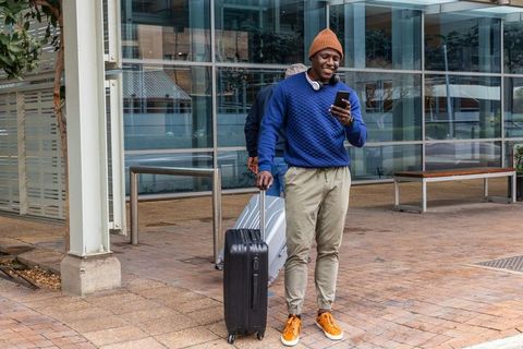 African american man walking with luggage checking smartphone outside glass building
