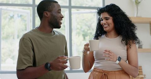 Black man and south asian woman sharing coffee, smiling and chatting in sunlit kitchen