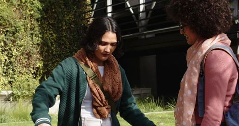 Two young women chatting on sunny college lawn wearing scarves and backpacks