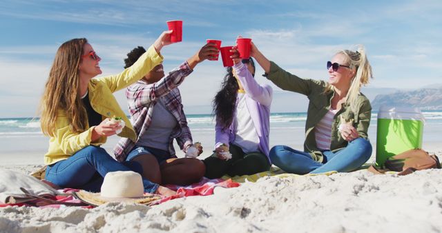 Group of diverse friends sitting on blanket enjoying picnic by sea. They are toasting with red cups, showing appreciation for their time together. Ideal for themes related to friendship, summer vacations, outdoor activities, and holiday celebrations.