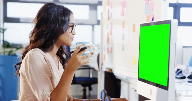Woman holding a coffee cup while working in a modern office. The computer monitor has a green screen, making it perfect for adding custom content. Ideal for use in articles about technology, productivity, remote work, and office environments.