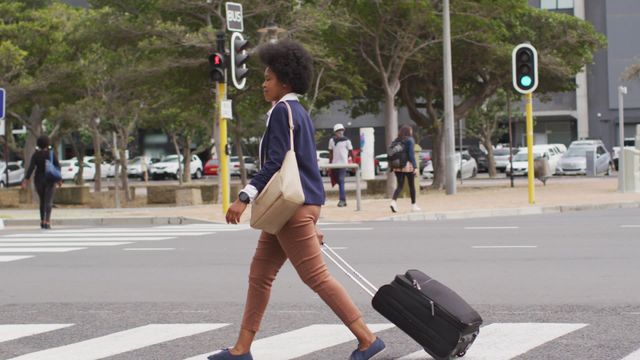 An African American businesswoman is walking across a city street while pulling a suitcase. She is dressed in a casual yet professional outfit, indicating a blend of business and comfort. Trees and other pedestrians can be seen in the background, showing a busy urban environment. This video can be used for topics related to business travel, professional lifestyle, digital nomadism, urban settings, and city life.