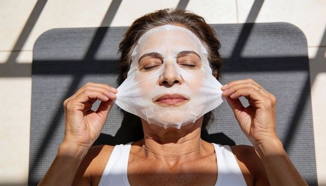 Senior woman removing sheet mask while relaxing on mat in sunlit minimalist interior