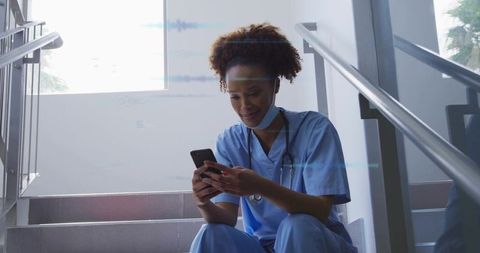Nurse sitting on hospital stairs checking smartphone during break with stethoscope and mask