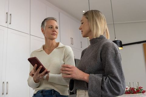 Adult daughter discussing smartphone with mature mother in bright modern kitchen