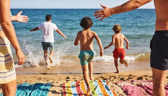Family running toward ocean on sunny beach with children in colorful swim trunks