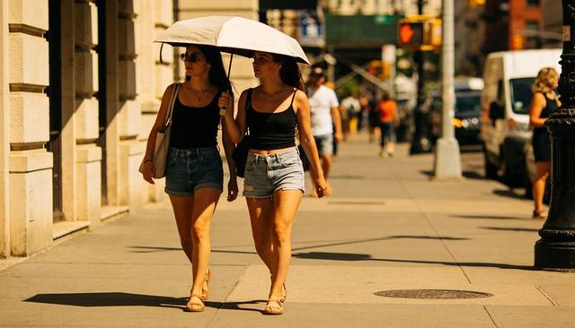Two young women walking under white umbrella on sunny city sidewalk in denim shorts and black tank t