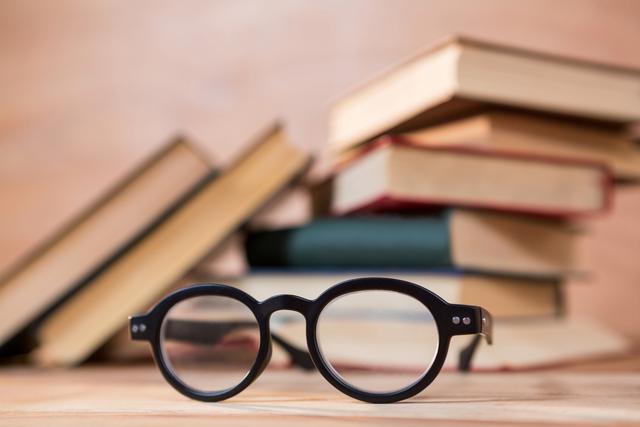 Close-up of spectacles resting on a wooden table with a stack of books in the background. Ideal for use in educational materials, library promotions, study guides, and articles related to reading, learning, and intellectual pursuits.