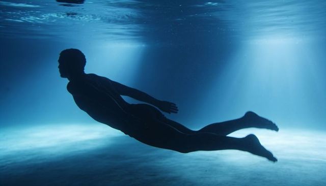 Gliding male silhouette wearing wetsuit under sunlit blue water over sandy seafloor
