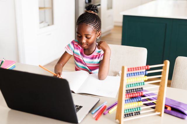 Young African American girl engaged in online learning at home, using a laptop and surrounded by school supplies. Ideal for illustrating concepts of remote education, home schooling, childhood learning, and the use of technology in education.