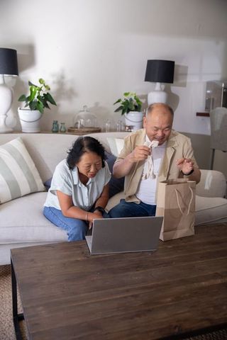 Senior asian couple unboxing online order while video calling on laptop in living room