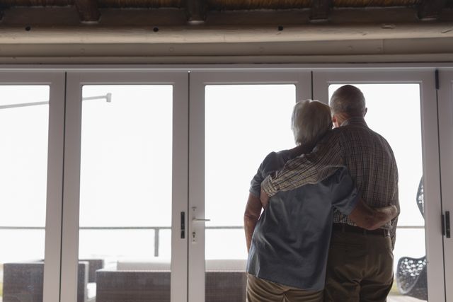 Rear view of active senior couple standing together while looking through the window at home