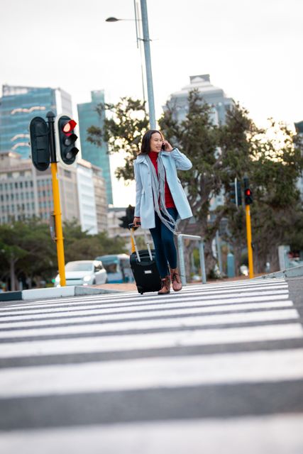 Asian woman crossing street with wheeling suitcase, talking on smartphone. Ideal for illustrating urban travel, commuting, business trips, and modern lifestyle themes. Suitable for use in travel blogs, business articles, and city life promotions.