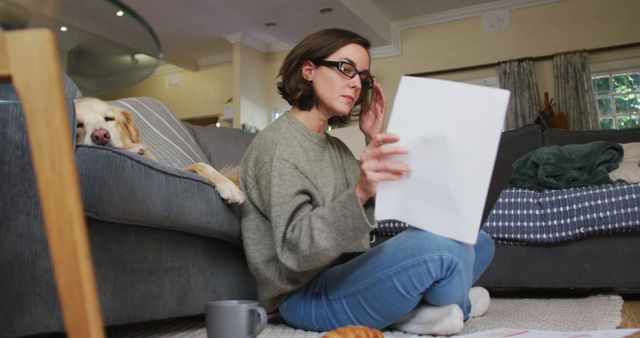 Woman sitting on floor in living room, reading a document while adjusting glasses. Dog is lying on sofa in the background. Scene includes a coffee cup and a croissant. Ideal for concepts related to remote work, relaxation, home life, multitasking, and comfort.