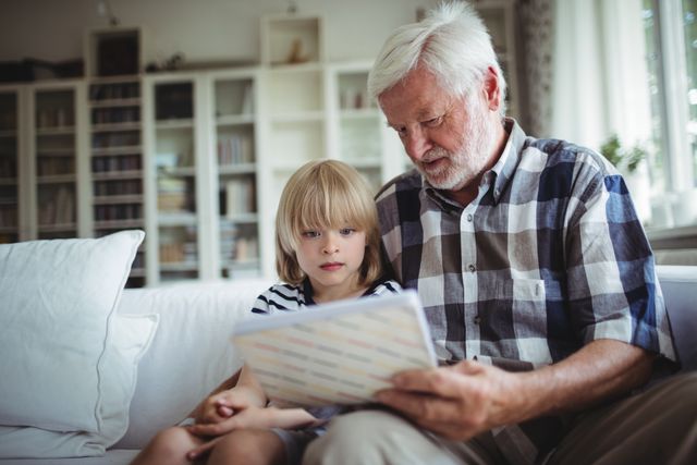 Grandfather and granddaughter sitting on couch looking at photo album. Perfect for themes of family bonding, intergenerational relationships, and nostalgic moments. Ideal for use in articles, advertisements, and social media posts about family, memories, and home life.