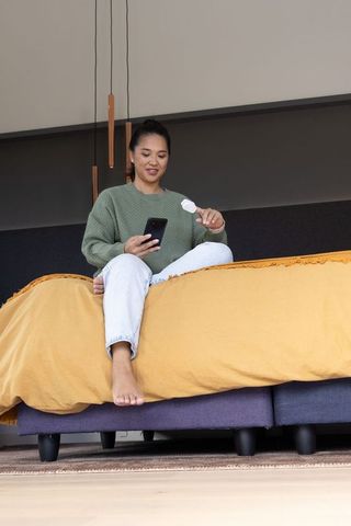 Asian woman sitting on bed edge holding smartphone and earbud case in modern bedroom