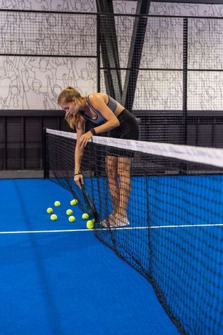 Female athlete scooping tennis balls over padel net on blue court wearing fitness watch