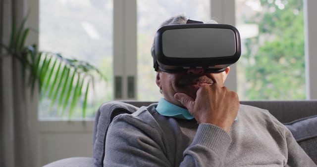 A joyful biracial senior man is engaging with virtual reality through a VR headset while relaxing at home. His genuine smile and expressive gestures suggest he is thoroughly enjoying the immersive experience. Ideal for use in articles and advertisements focusing on elderly well-being, modern technology impact on senior lifestyles, and home entertainment solutions. Can inspire more sales in VR devices tailored for older demographics and depict technology integration in retirement living.