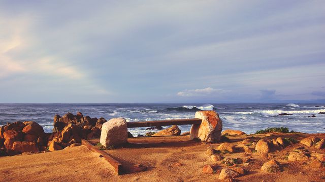 Rustic wooden bench positioned at rocky beach looking out to ocean waves during sunset. Ideal for content related to relaxation, travel inspirations, nature retreats, and coastal living. Perfect visual for articles on serene escapes, meditation spots, and oceanic views.