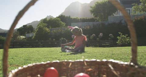 Young girl wearing bunny ears sitting on sunlit backyard lawn holding easter basket