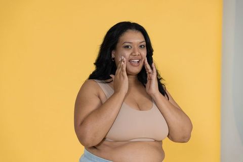Confident plus-size woman touching face wearing beige crop top, hoop earrings, yellow background