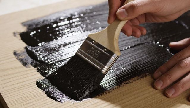 Applying glossy black wood stain with brush on pale timber plank close-up