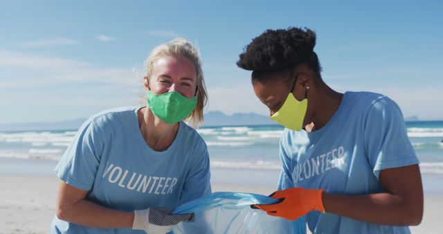 Two diverse women wearing volunteer t shirts and face masks picking up rubbish from beach. eco conservation volunteers, beach clean-up during coronavirus covid 19 pandemic.