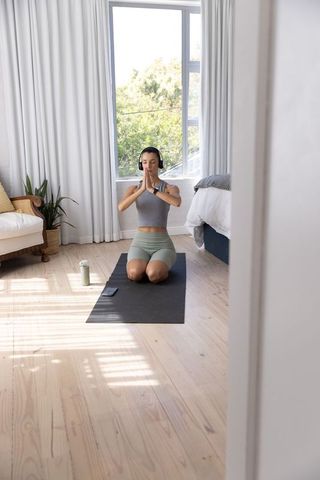 Woman practicing meditation on yoga mat in sunlit minimal bedroom wearing headphones