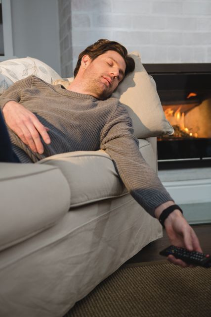 Man sleeping on sofa with TV remote in hand in cozy living room. Ideal for concepts of relaxation, leisure, home comfort, and evening routines. Suitable for lifestyle blogs, home decor advertisements, and articles on relaxation techniques.