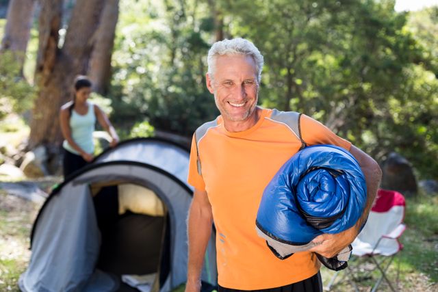 Mature man smiling and holding a sleeping bag at a campsite with a tent and trees in the background. Ideal for use in advertisements, travel blogs, camping gear promotions, and outdoor adventure articles.