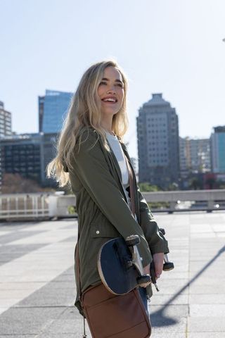 Young woman smiling and holding skateboard on rooftop promenade with urban skyline