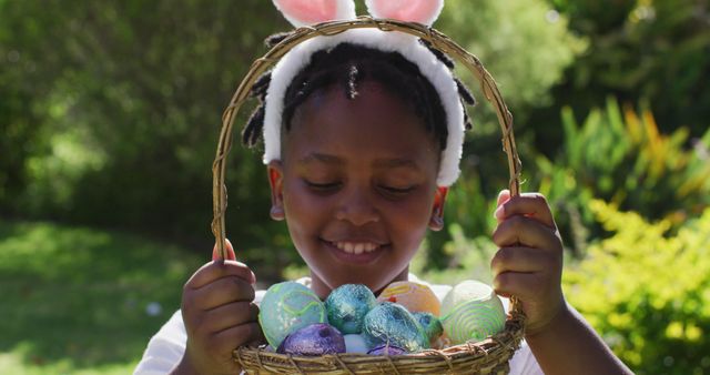 Smiling african american girl wearing easter bunny ears holding basket of easter eggs in garden. family spending easter time together staying at home in isolation during quarantine lockdown.