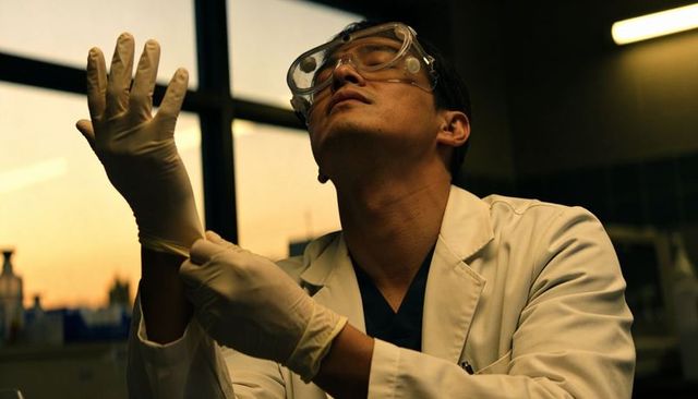 Asian scientist pulling on gloves at lab bench near sunset window, lab coat and goggles