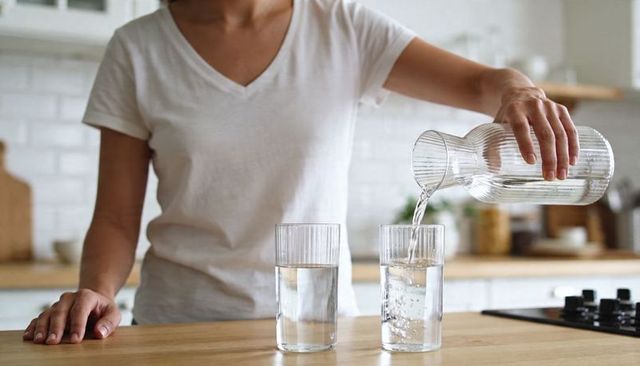 Pouring water from ribbed glass pitcher into tall glass on wooden kitchen countertop
