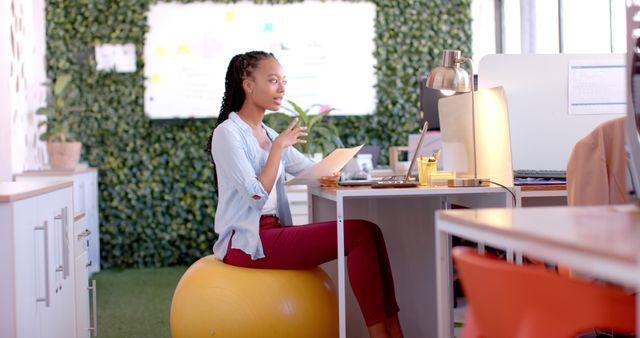 Young woman interacting with a digital device while seated on a yellow exercise ball in a contemporary office. This photo is ideal for illustrating flexible working environments, modern workspaces, ergonomic solutions, and productivity in professional settings. Great for articles and advertisements about workplace wellness, office design, and technology use in offices.